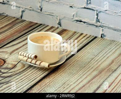 Una tazza di cappuccino fresco su un piattino con due involtini di cialda su un tavolo di legno chiaro. Primo piano. Foto Stock