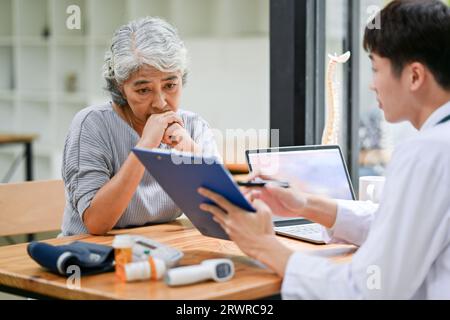 Una vecchia signora asiatica in pensione stressata e seria sta consultando i piani di trattamento dopo l'intervento chirurgico con un medico in una sala d'esame in un ospedale. Foto Stock