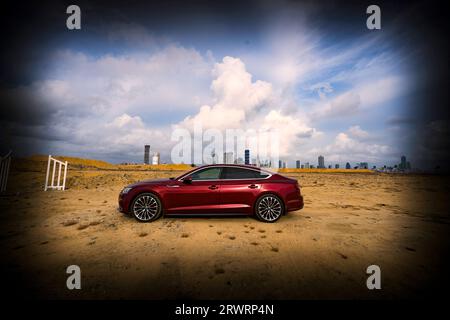 Una vettura sportiva Audi A5 rossa parcheggiata su un terreno sabbioso con un paesaggio urbano sullo sfondo Foto Stock