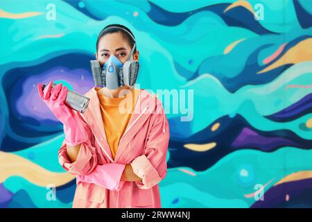 Graffiti artista di strada donna con maschera respiratoria in piedi vicino al muro con i suoi dipinti che guardano alla fotocamera con spazio per la copia. Concetto di arte urbana Foto Stock