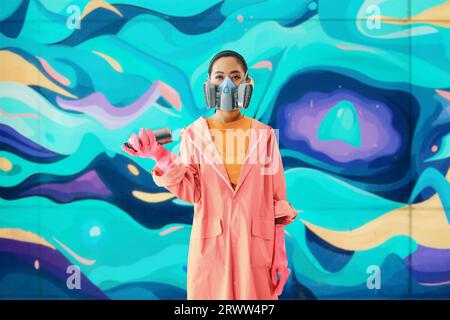 Ritratto di artista di strada femminile in maschera respiratoria in piedi vicino alla parete dei graffiti con i suoi dipinti che guardano alla macchina fotografica. Concetto di arte urbana Foto Stock