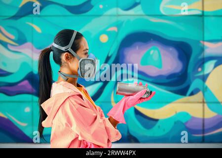 Vista del profilo di una Street artist femminile con maschera respiratoria sullo sfondo della parete con i suoi dipinti colorati con spazio per la copia. Concetto di arte urbana Foto Stock