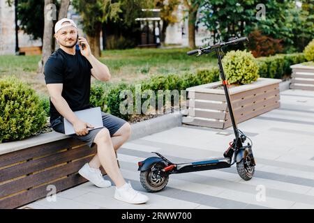 Un bel freelance che tiene un computer portatile e un telefono parlante seduto sulla panca vicino a uno scooter elettrico per strada Foto Stock