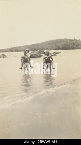 Alfred Tamlin e un altro uomo europeo, entrambi con elmetti di piombo, sono trasportati in acque poco profonde sul dorso di due uomini africani che fumano sigarette. Sono vicini alla spiaggia e gli alberi sono visibili sulle colline dietro di loro. Foto Stock