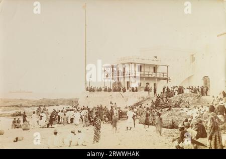 Vista di un grande edificio ufficiale imbiancato, forse una dogana, sulla riva di Cape Coast. C'è un gran numero di persone sul balcone dell'edificio e la folla che si fregia intorno alla riva sottostante. Alcune persone sono in uniforme, forse poliziotti o doganieri. La SS Karina può essere vista in mare sullo sfondo, una nave silurata al largo della costa di Irelend il 1° agosto 1917. Didascalia del testo originale: Cape Coast. Gold Coast. 1917 mostra S.S. 'Karinaa' (silurata 1/ 1917 agosto). Foto Stock