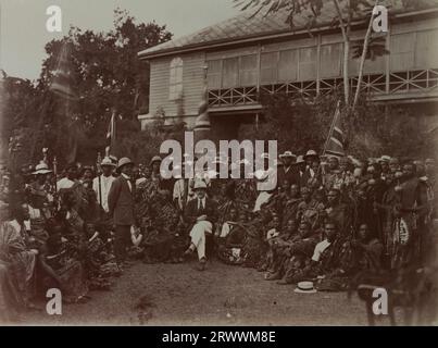 Un uomo europeo che indossa un elmetto con stemma e posa per un ritratto di gruppo con i capi Asante fuori da un grande edificio in stile coloniale. Alcuni degli Asante indossano le legature Kente e tengono in mano il personale, e molti portano bandiere Union Jack. Foto Stock