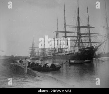 Una fila di barche sampan è ormeggiata su un'insenatura del fiume con grandi navi a vela dietro. Un uomo europeo che indossa un vestito leggero e un casco sta camminando sulla riva del fiume. Foto Stock