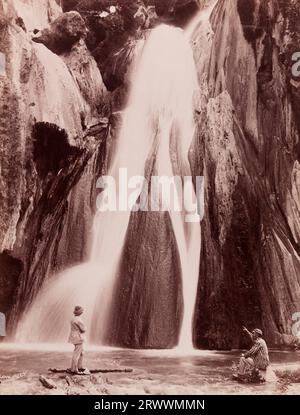 Vista di una cascata su una ripida parete rocciosa. Due uomini europei sono alla base della piscina, uno vestito formalmente con un casco e l'altro con un cappello a righe e un blazer che puntano verso la cascata. Scritto in negativo: Lawrie & Co. Foto Stock