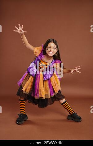 Ragazza spaventosa in costume da strega di Halloween che ringhia e gesti su sfondo marrone, per tutta la lunghezza Foto Stock