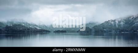 Panorama su un lago blu riflettente e una montagna limpida boscosa con foreste di abeti rossi di conifere sempreverdi contro il cielo nuvoloso blu diurno della Norvegia Foto Stock