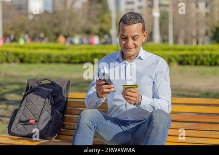 Un giovane latinoamericano che fa un acquisto con una carta di credito sul suo cellulare all'aperto. Foto Stock