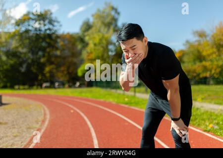 Giovane asiatico che fa sport allo stadio. Si trova piegato sul tapis roulant, si copre la bocca con la mano, si sente molto nauseato. Foto Stock