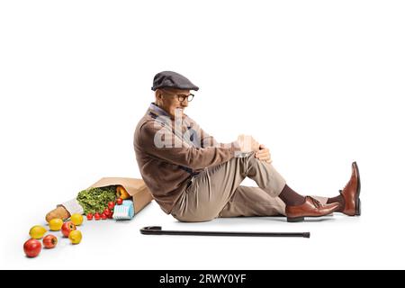 Uomo anziano con una borsa di roceria seduto sul pavimento e tenendo la gamba isolata su sfondo bianco Foto Stock