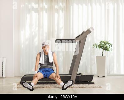 Anziano stanco seduto su un tapis roulant a casa e sorridente Foto Stock