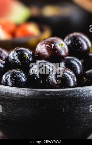 Jaboticaba o jabuticaba, frutti brasiliani e sudamericani, in una pentola di argilla con frutti tropicali sullo sfondo. Foto Stock