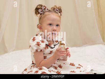 Una bella bambina che indossa un apparecchio acustico siede sul letto con un bel vestito e gioca con il profumo della madre. Annusa gli aromi Foto Stock