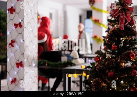 Albero di Natale decorato per festività da vicino con uomini d'affari che lavorano in uno sfondo sfocato. Ufficio moderno con graziose luci di Natale che decorano le sale durante la stagione delle vacanze invernali Foto Stock