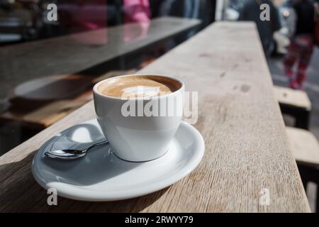 Messa a fuoco selezionata e vista ravvicinata di una tazza di cappuccino in ceramica bianca sulla parte superiore con una bella latte art delicata con motivo a cuore, su legno all'aperto. Foto Stock