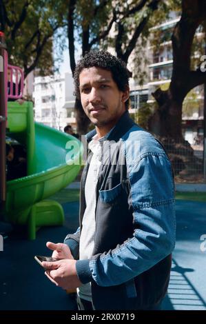 immagine verticale di un uomo venezuelano afro-latino che indossa una giacca jean blu, che tiene il telefono in piedi nel parco guardando la fotocamera sorridendo leggermente. Foto Stock