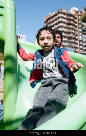 immagine verticale, bambino maschio seduto nel parco giochi pubblico felice di guardare la macchina fotografica con suo padre dietro di lui, concetto di stile di vita. Foto Stock