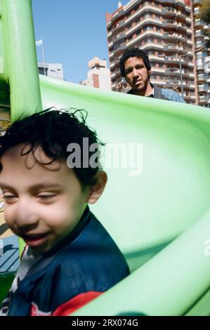 immagine verticale, bambino maschio seduto nel parco giochi pubblico felice di guardare la macchina fotografica con suo padre dietro di lui, concetto di stile di vita. Foto Stock