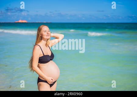 Radiosa donna incinta in costume da bagno, sullo sfondo mozzafiato di un mare turchese. Bellezza serena della maternità vicino alla riva Foto Stock