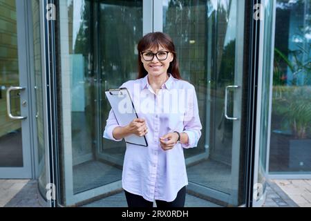 Ritratto di una donna d'affari di mezza età che tiene un appunti vicino alle porte di uffici in vetro Foto Stock