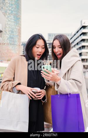Ritratto verticale di giovani donne felicemente multietniche sorprese delle vendite e offre un telefono cellulare che fa acquisti online. Signore e amici che acquistano vestiti utilizzando lo smartphone. Foto di alta qualità Foto Stock