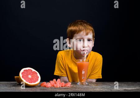 il ragazzo beve succo amaro e aspro dal pompelmo rosso, il succo non è delizioso e il bambino non piace, utile ma non delizioso grappa Foto Stock