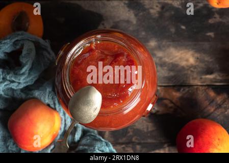 Deliziosa marmellata di albicocche fatta in casa in un barattolo di vetro su un tavolo di legno. C'è anche un piccolo cucchiaio di metallo, frutti di albicocca interi, metà frutta. Vista dall'alto Foto Stock