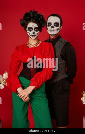 coppia in abiti festivi e trucco catrina in posa su sfondo rosso, festa dia de los muertos Foto Stock