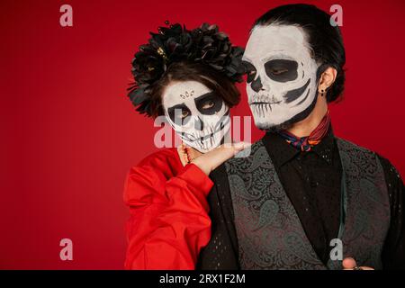 Donna con trucco catrina e corona nera appoggiata a spalla di un uomo inquietante sul rosso, Day of Dead Foto Stock