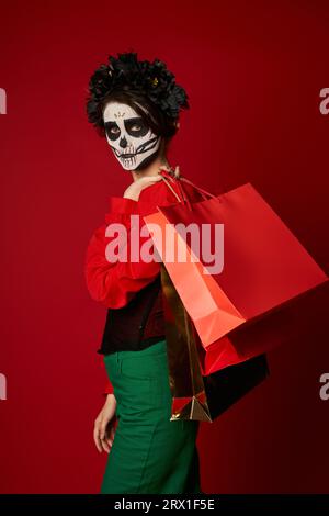 donna elegante con corona nera e trucco catrina che tiene le borse della spesa e guarda la fotocamera sul rosso Foto Stock