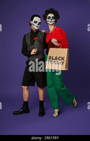 donna in inquietante trucco catrina in posa con la borsa della spesa del black friday vicino a un uomo spettrale su blu Foto Stock