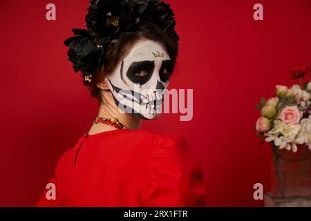 ritratto di una giovane donna con lo spaventoso trucco catrina e corona nera che guarda la macchina fotografica su rosso Foto Stock