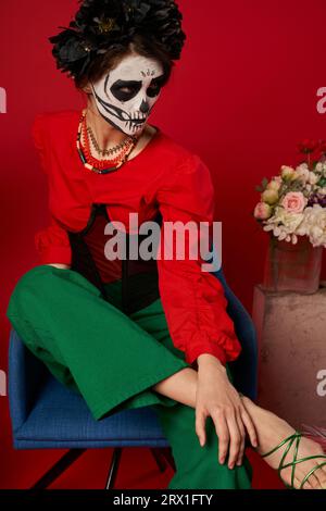 donna con trucco catrina seduta in poltrona vicino ai fiori sul rosso, tradizione dia de los muertos Foto Stock