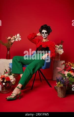 makeup donna in dia de los muertos seduta in poltrona vicino all'altare tradizionale con fiori rossi Foto Stock