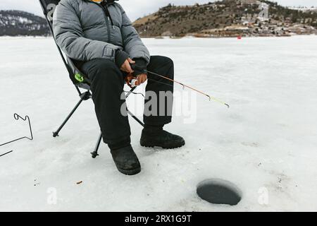 Il ragazzo si siede sul lago ghiacciato per pescare in un buco di ghiaccio in inverno in cappotto Foto Stock