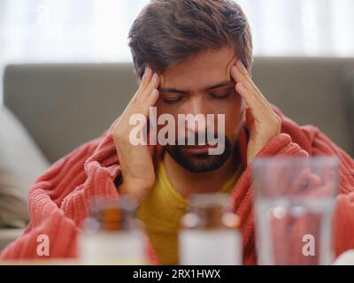 stagione autunnale e invernale degli starnuti. Giovane uomo caucasico con influenza si sente freddo in salotto a casa, avendo febbre e mal di testa e toccando la fronte per controllare la sua temperatura. Foto Stock