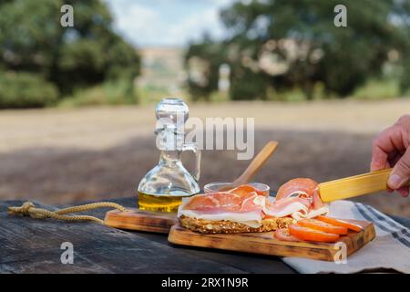 Donna che mette fette di prosciutto iberico alimentato con ghianda su una fetta di pane con pomodoro e olio d'oliva su un tavolo di legno in campagna, tipico spagnolo Foto Stock