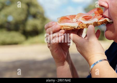 Donna di profilo che mangia un brindisi di pane con pomodoro e prosciutto iberico alimentato a ghiande in campagna con una foresta sullo sfondo e un cielo blu con Foto Stock