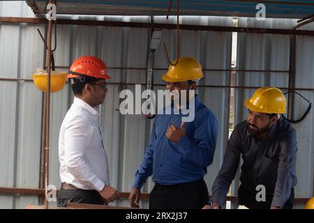 Tre ingegneri edili maschili in abbigliamento e cappelli rigidi discutono di un progetto sotto un riparo presso il sito Foto Stock