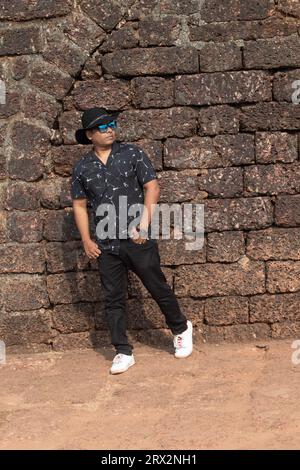 Bell'uomo con cappello da cowboy contro un antico muro di pietra. Elegante viaggiatore maschile con cappello e occhiali da sole estivi presso il sito storico Foto Stock