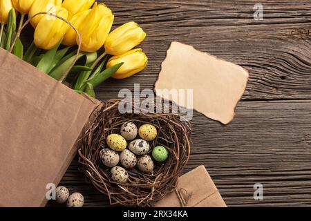 Tulipani gialli in un sacchetto di carta, un nido con le uova di Pasqua su uno sfondo di legno. Vista da sopra con copia spazio. Foto Stock