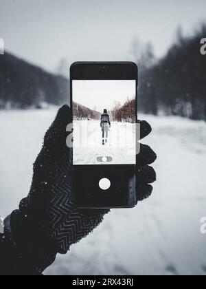 Uomo con guanti invernali che tiene lo smartphone e scatta una foto (vista pov) di una donna che cammina sulle montagne innevate. Foto Stock