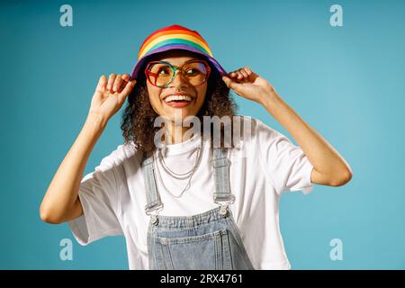 La donna sorridente con occhiali da vista indossa un berretto arcobaleno mentre guarda lontano sullo sfondo blu dello studio Foto Stock