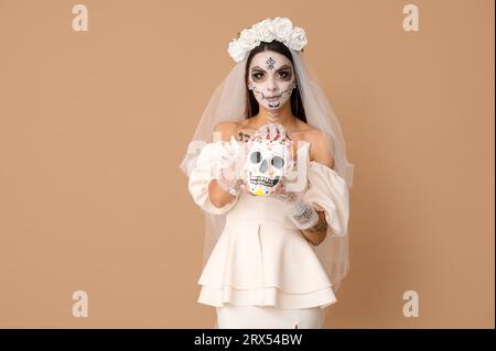 Giovane donna vestita da sposa morta per la festa di Halloween con teschio umano dipinto su sfondo beige Foto Stock