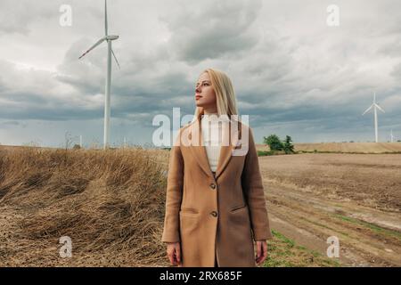 Donna bionda in piedi davanti alla turbina eolica sotto il cielo nuvoloso Foto Stock