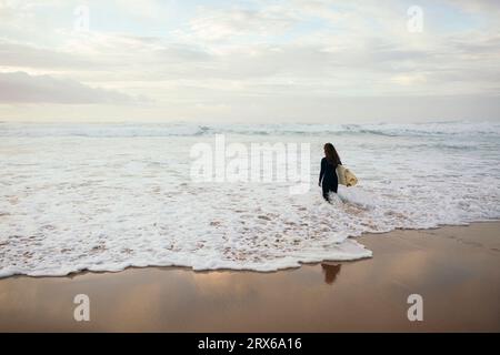 Giovane surfista con tavola da surf che cammina in mare ondulato in spiaggia Foto Stock