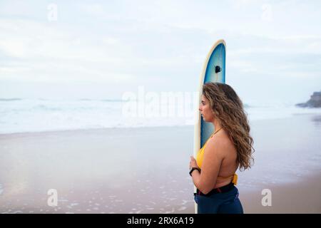 Donna premurosa con tavola da surf che guarda il mare sulla spiaggia Foto Stock